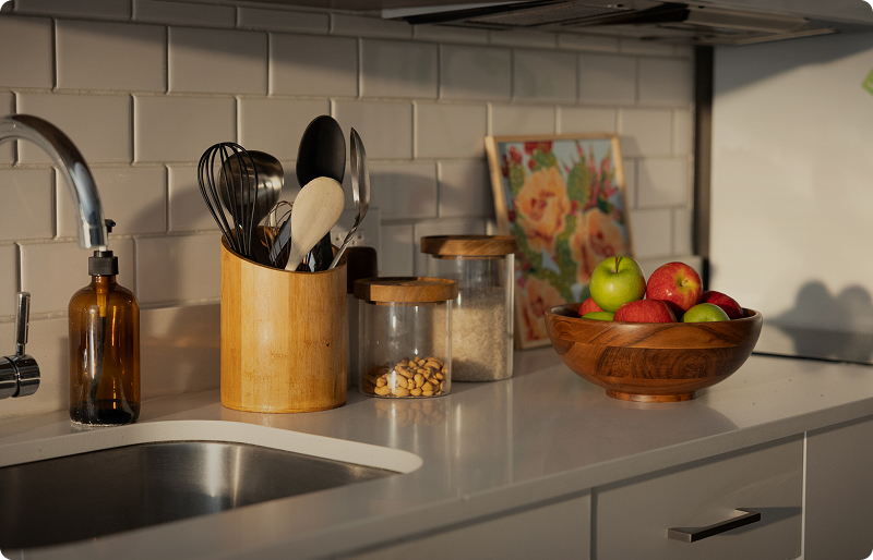 kitchen countertop with a utensil holder, glass food jars and a wooden bowl filled with apples